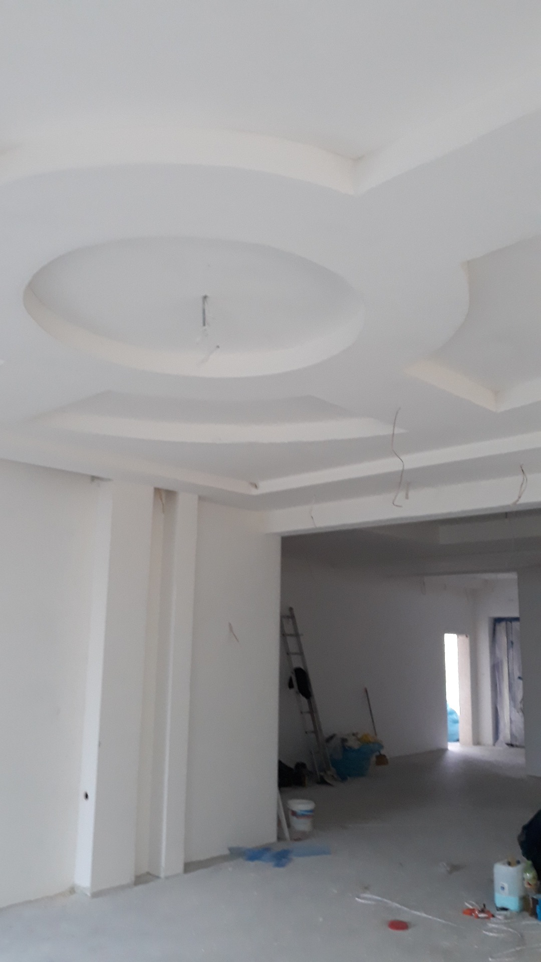 Białe, nowo wykończone wnętrze z dekoracyjnym sufitem podwieszanym z okrągłym ornamentem, widoczna drabina, narzędzia i wiadro z farbą, w tle otwarte przejście do kolejnego pomieszczenia.