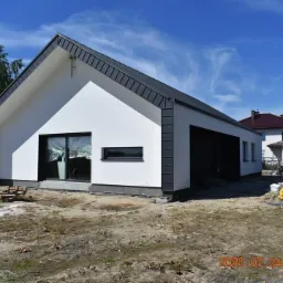 Nowoczesny, parterowy dom z białymi ścianami i ciemnoszarą stolarką okienną oraz dachem, widoczny w słoneczny dzień na tle błękitnego nieba z lekkimi chmurami, otoczony niezagospodarowanym terenem.