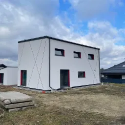 Nowoczesny, biały dom z elementami dekoracyjnymi w postaci czarnych linii i czerwonych obramowań okien, widoczny w surowym otoczeniu placu budowy z betonowymi płytami i innymi budynkami w tle.