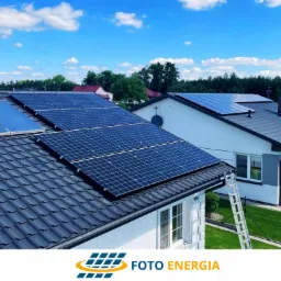 Panele słoneczne na dachu domu jednorodzinnego, widoczna drabina oparta o rynnę i logo firmy Foto Energia w dolnej części zdjęcia.