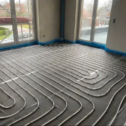 Białe rury ogrzewania podłogowego ułożone spiralnie na czarnej macie w surowym pomieszczeniu z dużymi oknami, zabezpieczonymi folią ochronną.