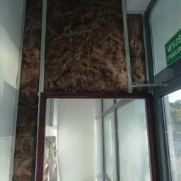 Wnętrze budynku w trakcie prac: widoczna wełna mineralna w metalowym stelażu, przygotowana pod montaż płyt kartonowo-gipsowych, obok okno z napisem 'Wyjście Ewakuacyjne' i materiały budowlane.