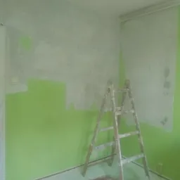 Pokój w trakcie remontu: ściany częściowo pomalowane na zielono i szpachlowane, drewniana drabina stoi na podłodze pokrytej resztkami gładzi, widoczne niedoskonałości malowania przy suficie.