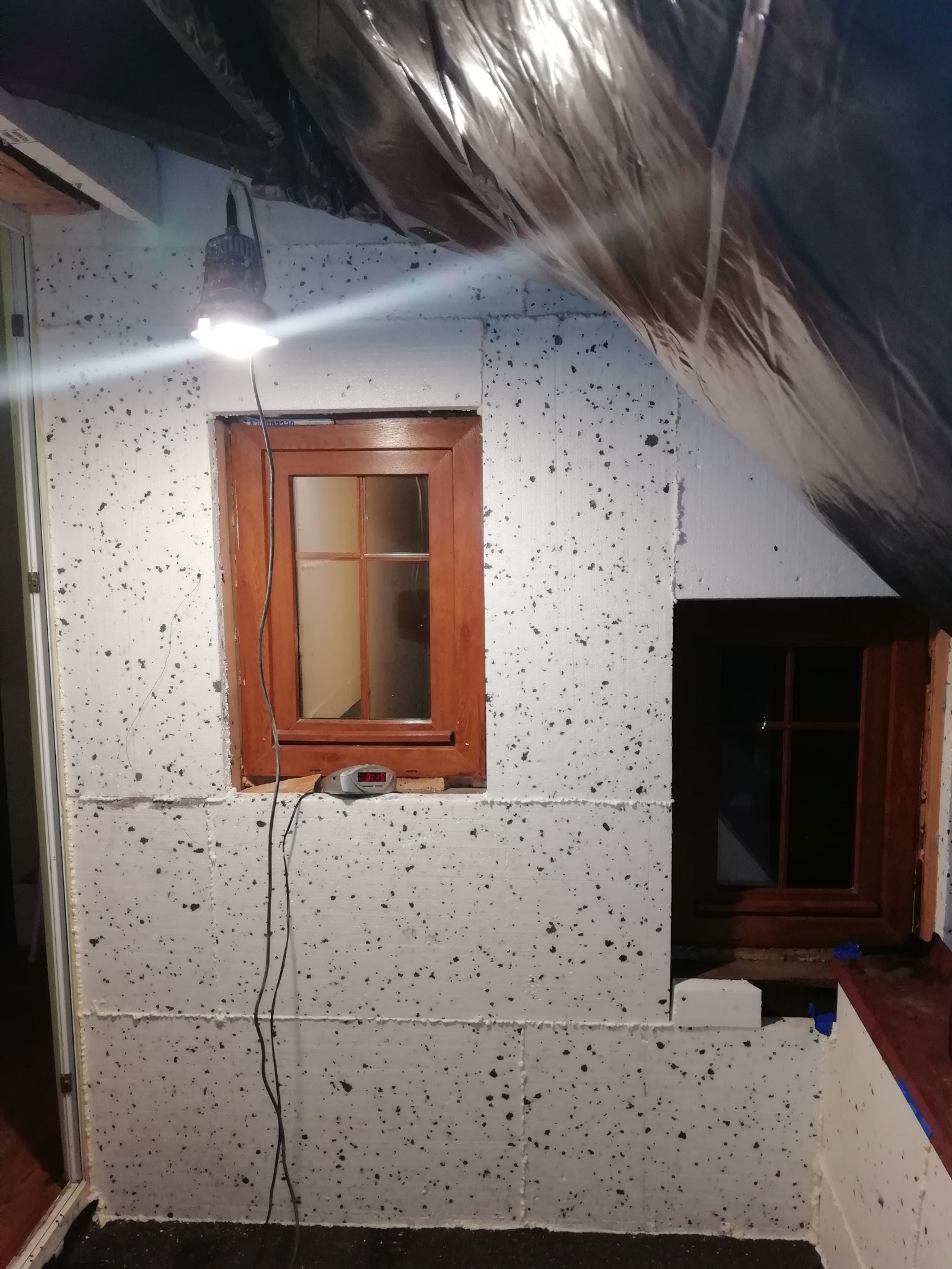 Wnętrze poddasza w trakcie prac izolacyjnych: ściany wyłożone styropianem z widocznymi połączeniami, drewniane okna, kabel od lampy roboczej zwisający wzdłuż ściany, czarna folia zabezpieczająca...