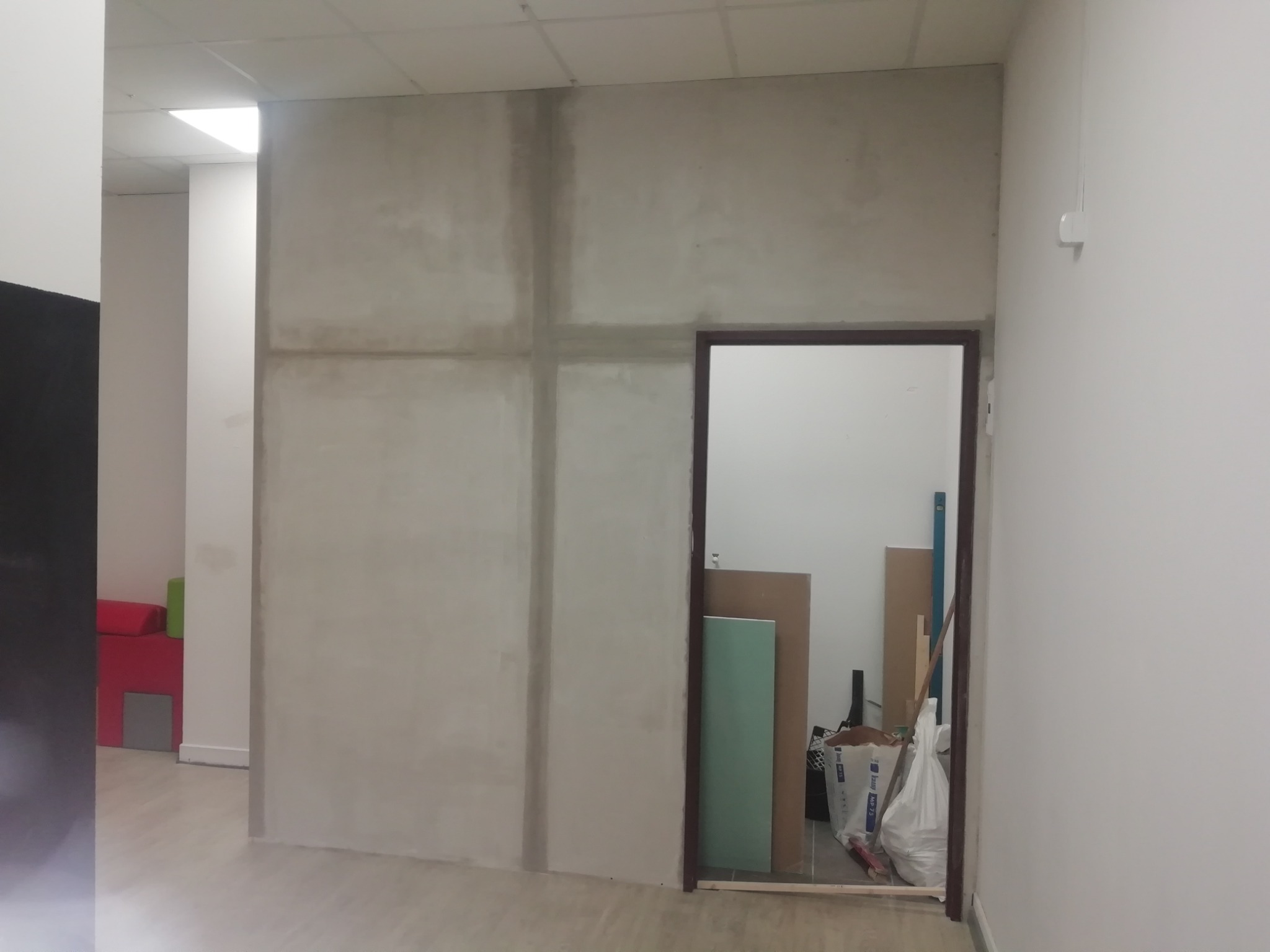 Ściana z płyt kartonowo-gipsowych w trakcie szpachlowania, widoczne łączenia płyt, w tle otwarte drzwi z materiałami budowlanymi.