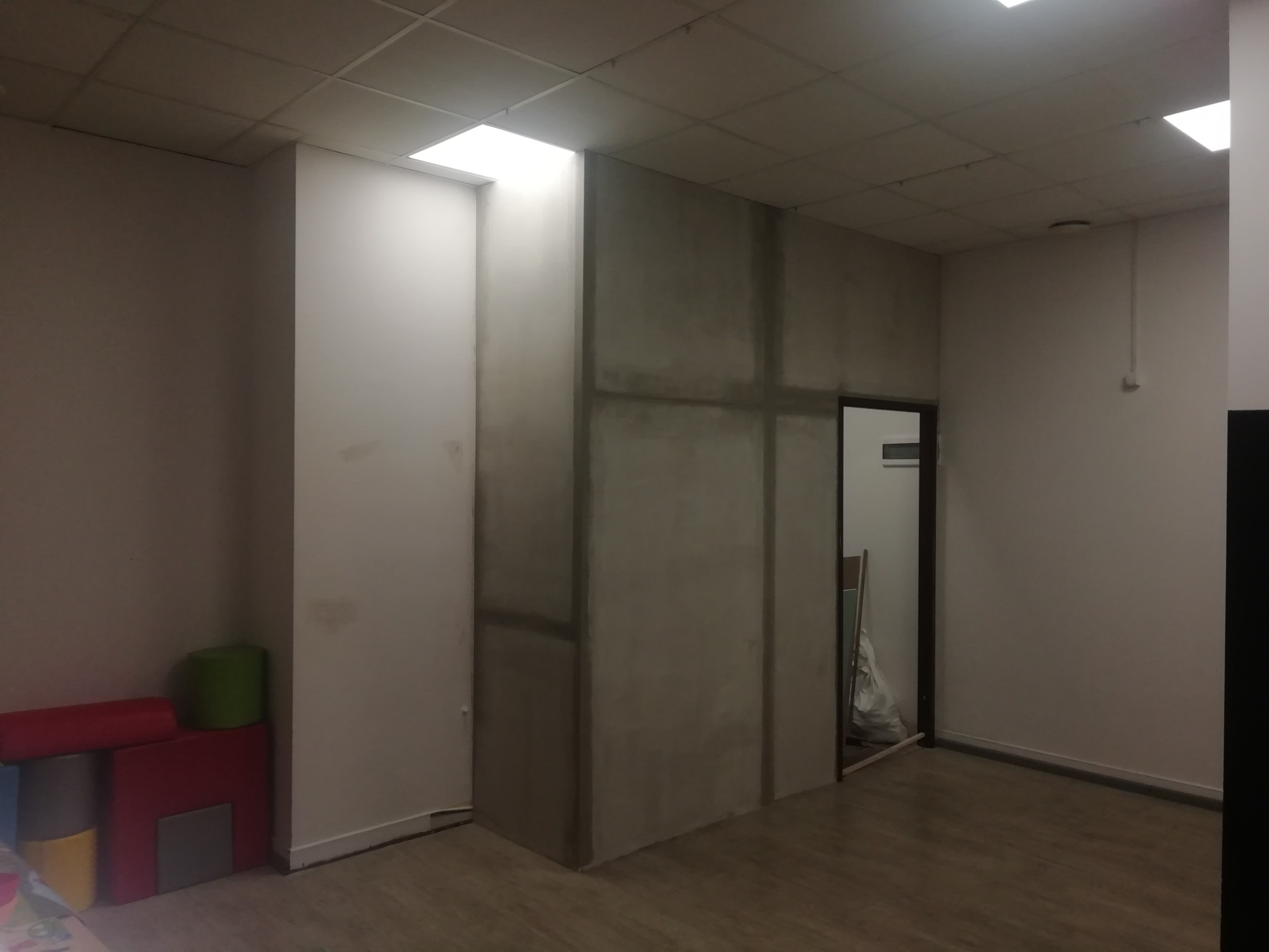 Wnętrze w trakcie prac wykończeniowych, surowa ściana z płyt kartonowo-gipsowych z widocznymi łączeniami, drewnopodobna podłoga, widoczne elementy instalacji pod sufitem.