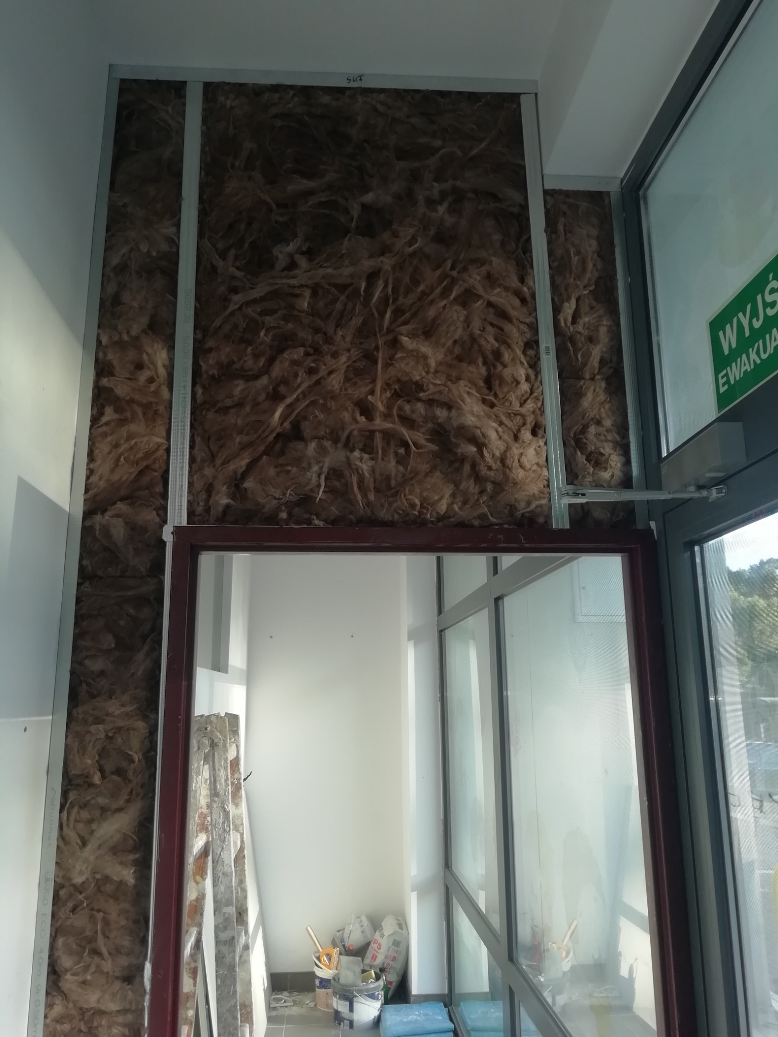 Wnętrze budynku w trakcie prac: widoczna wełna mineralna w metalowym stelażu, przygotowana pod montaż płyt kartonowo-gipsowych, obok okno z napisem 'Wyjście Ewakuacyjne' i materiały budowlane.