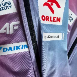 Zbliżenie na kurtkę sportową z naszytymi logotypami sponsorów, w odcieniach fioletu i błękitu, z widocznymi detalami szwów i zamka błyskawicznego.