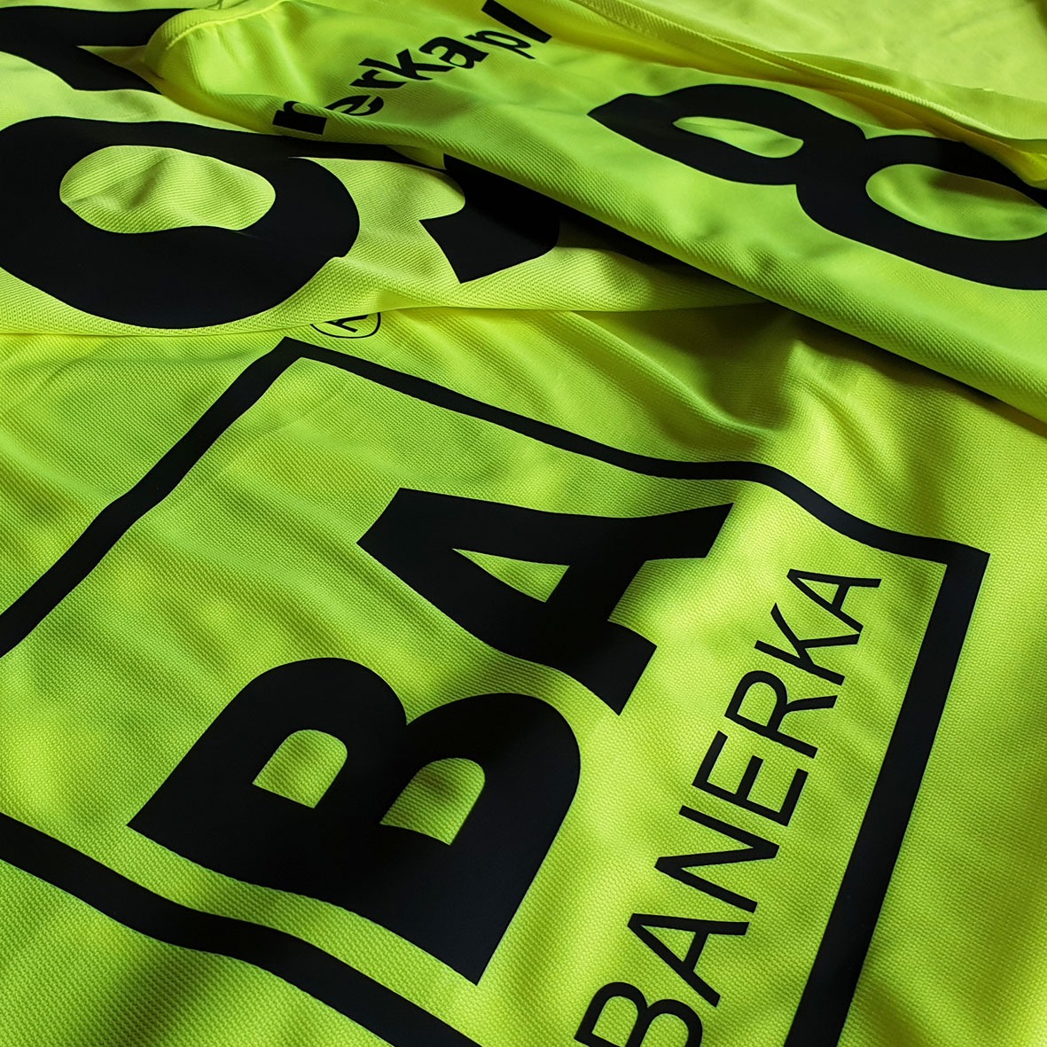 Jaskrawożółte koszulki sportowe z czarnym nadrukiem 'BANERKA' i numerem '8', ułożone warstwami, widoczna faktura materiału.