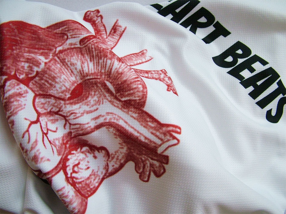 Biała koszulka z nadrukiem anatomicznego serca w kolorze czerwonym i napisem 'Heart Beats' w kolorze czarnym, widoczne detale faktury materiału.