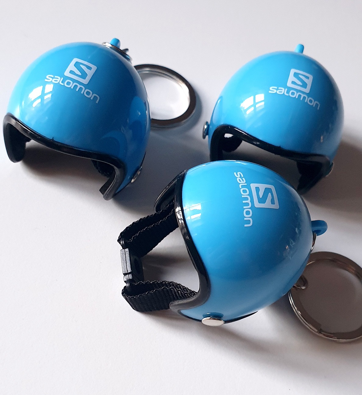 Trzy błękitne, plastikowe kaski w miniaturze z logo Salomon, jeden z czarnym paskiem, leżące na białym tle.
