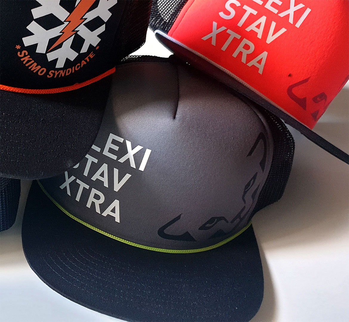 Stos czapek z daszkiem: czarna z białym nadrukiem 'SKIMO SYNDICATE' i pomarańczowym sznurkiem, szara z białym napisem 'LEXI STAV XTRA' i zielonym sznurkiem, czerwona z białym nadrukiem 'LEXI STAV...