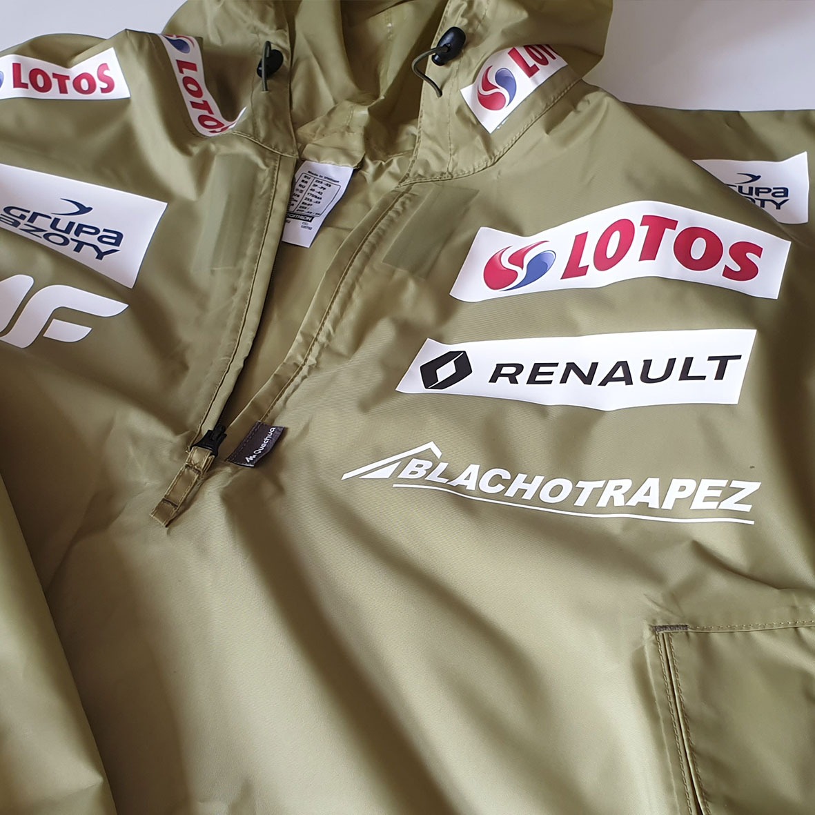 Oliwkowa kurtka sportowa z kapturem, pokryta licznymi nadrukami logo sponsorów, widoczny zamek błyskawiczny i metka.