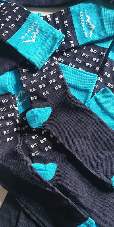 Stos ciemnych skarpet z turkusowymi palcami i piętami, ozdobionych powtarzającym się wzorem 'BS' oraz logo 'Livigno 20' z motywem górskim.