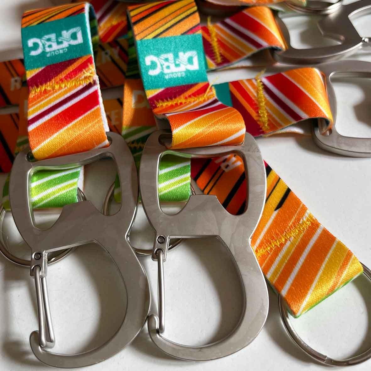 Metalowe otwieracze do butelek z kolorowymi, wzorzystymi paskami z logo DFBG Group, przymocowane do kółek, leżące na białym tle.