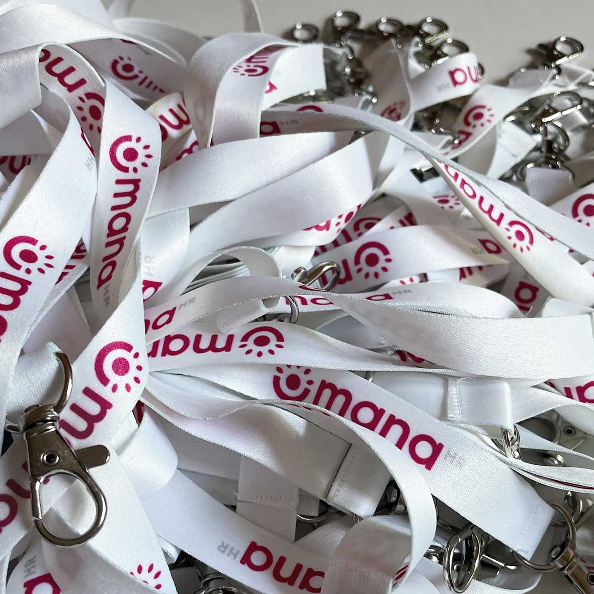 Stos białych smyczy z karabińczykami, z nadrukowanym czerwonym logo i nazwą firmy 'mana HR'.