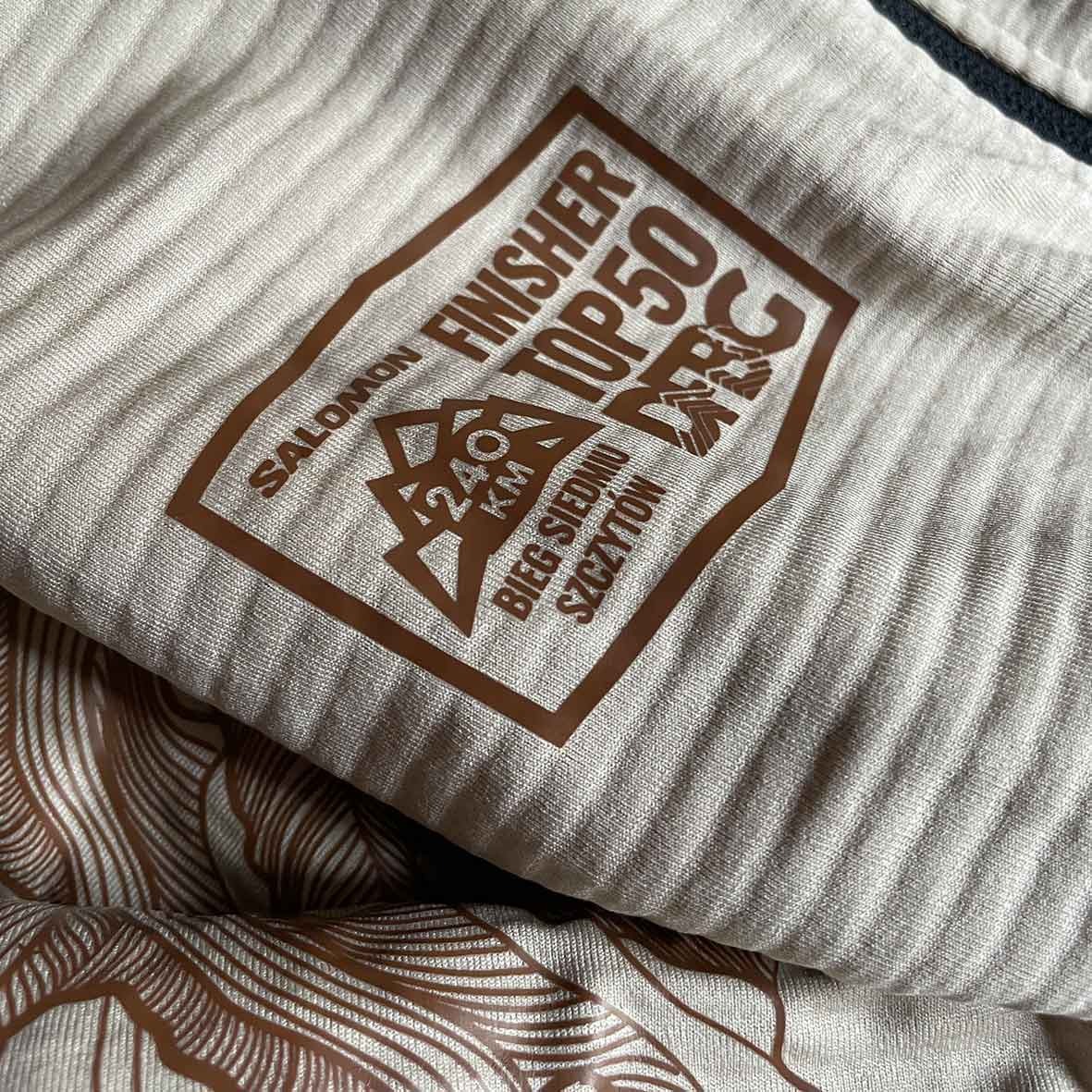 Beżowa koszulka z brązowym nadrukiem: logo Salomon, napis FINISHER TOP 50, 240 km Bieg Siedmiu Szczytów, widoczne delikatne prążki tkaniny i fragment drugiego nadruku z motywem górskim.