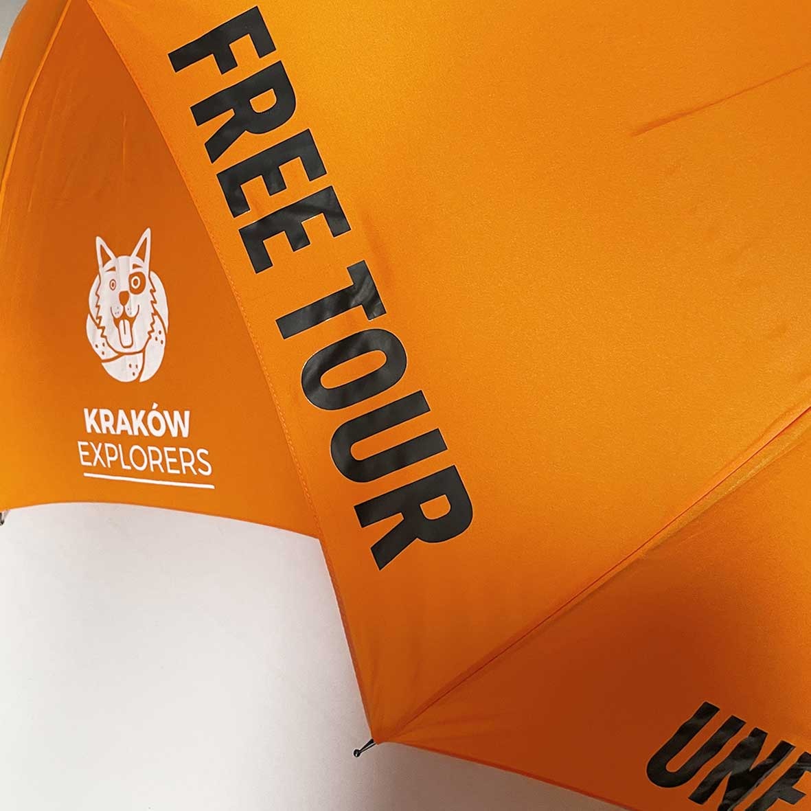 Pomarańczowy parasol z nadrukowanym logo 'Kraków Explorers' i napisem 'FREE TOUR', widoczne fragmenty konstrukcji parasola.
