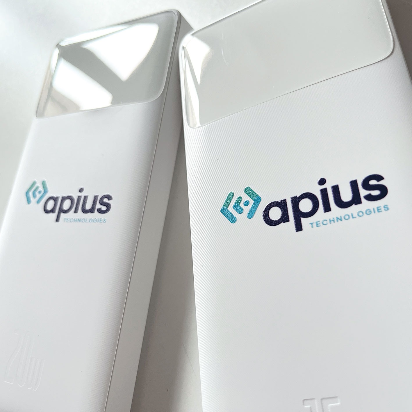 Dwa białe powerbanki z nadrukowanym logo 'apius TECHNOLOGIES' w kolorze niebiesko-czarnym, leżące na białym tle.