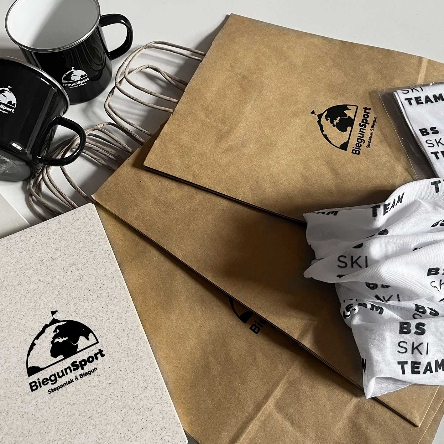 Zestaw promocyjny marki sportowej: czarne emaliowane kubki, papierowe torby z logo, notatnik i chusta wielofunkcyjna z nadrukiem 'BS SKI TEAM', ułożone na białym tle.
