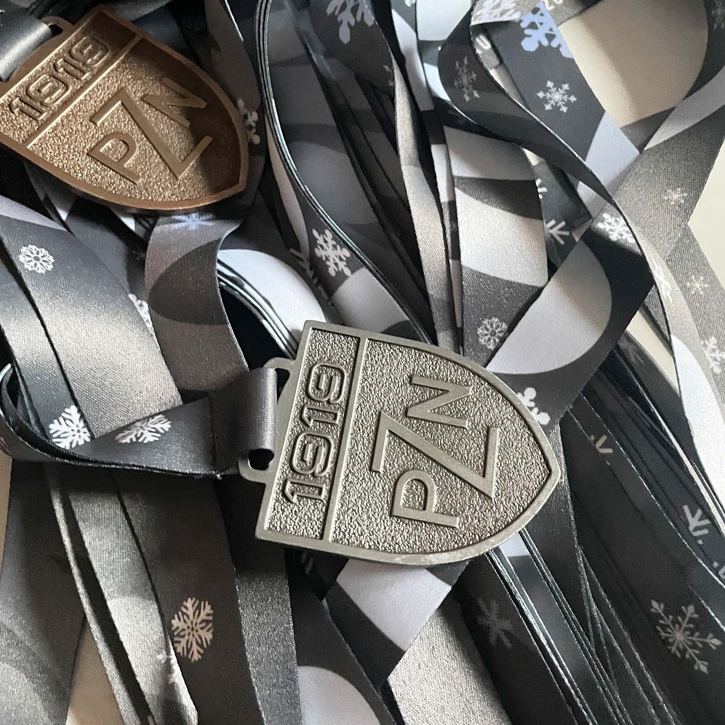 Dwie metalowe zawieszki w kształcie tarczy z napisem '1919 PZN' na tle pofałdowanych, czarno-białych wstążek z motywem śnieżynek.