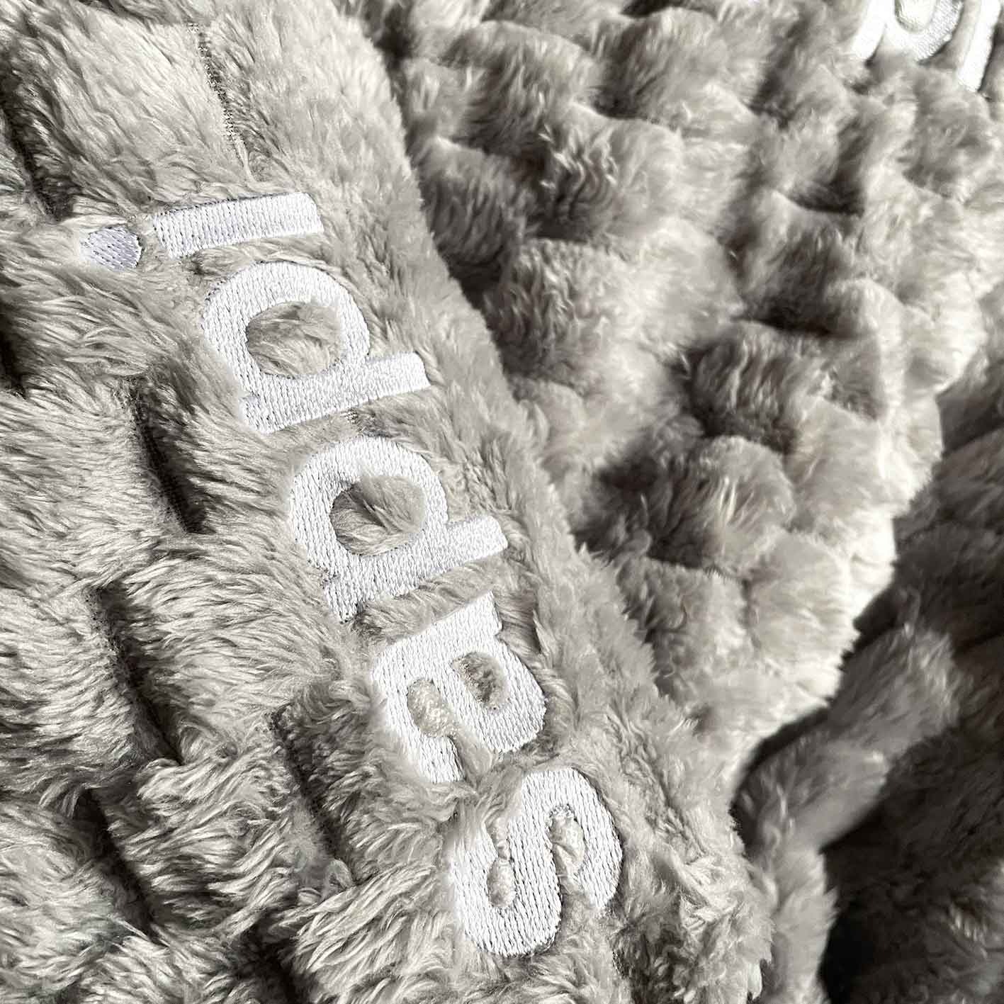 Szary, puszysty materiał z wytłoczonym wzorem i wyhaftowanym białym napisem, ujęcie z bliska, widoczne detale faktury.