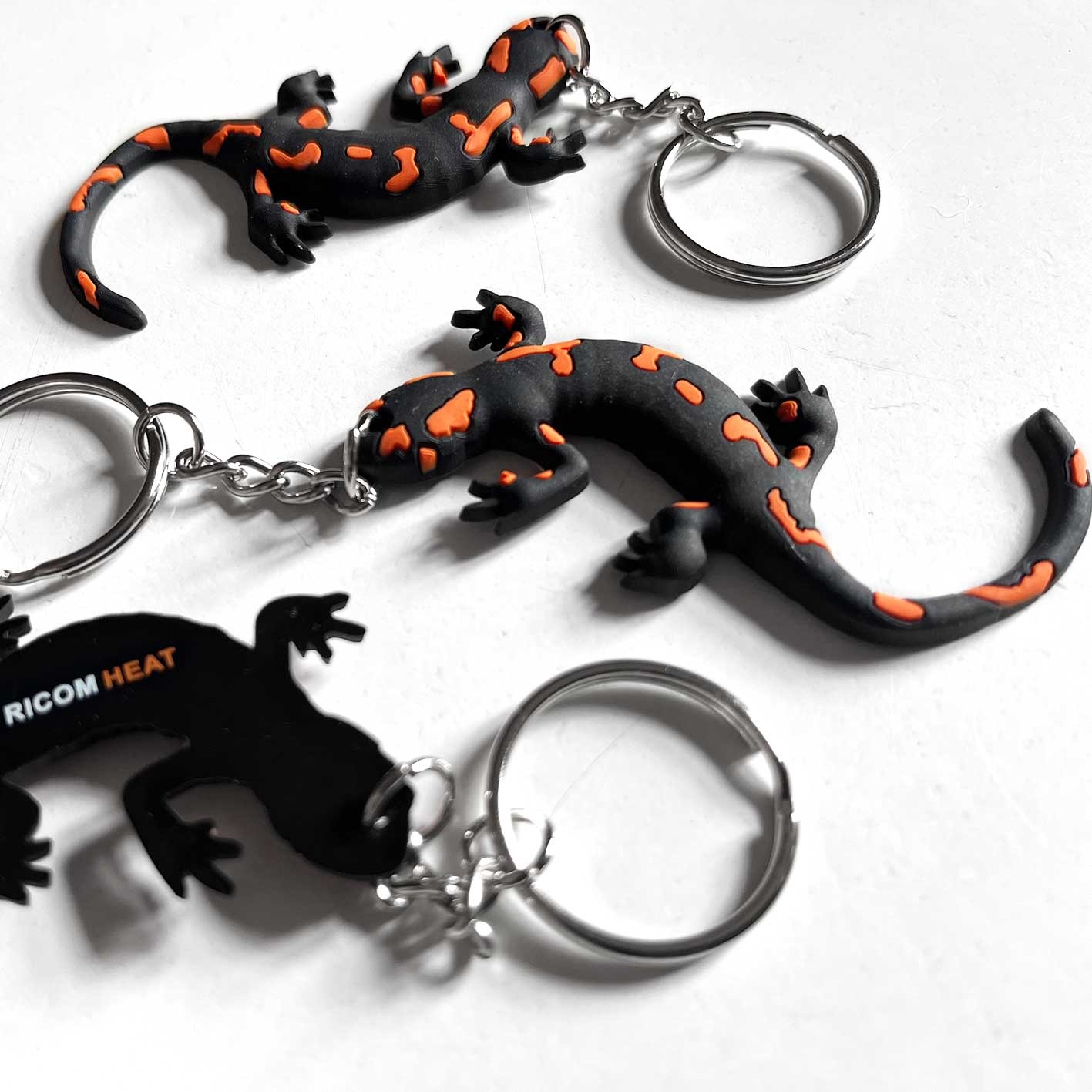Trzy breloczki w kształcie salamandry plamistej, dwa czarno-pomarańczowe i jeden czarny z napisem 'RICOM HEAT', przymocowane do metalowych kółek, leżą na białym tle.