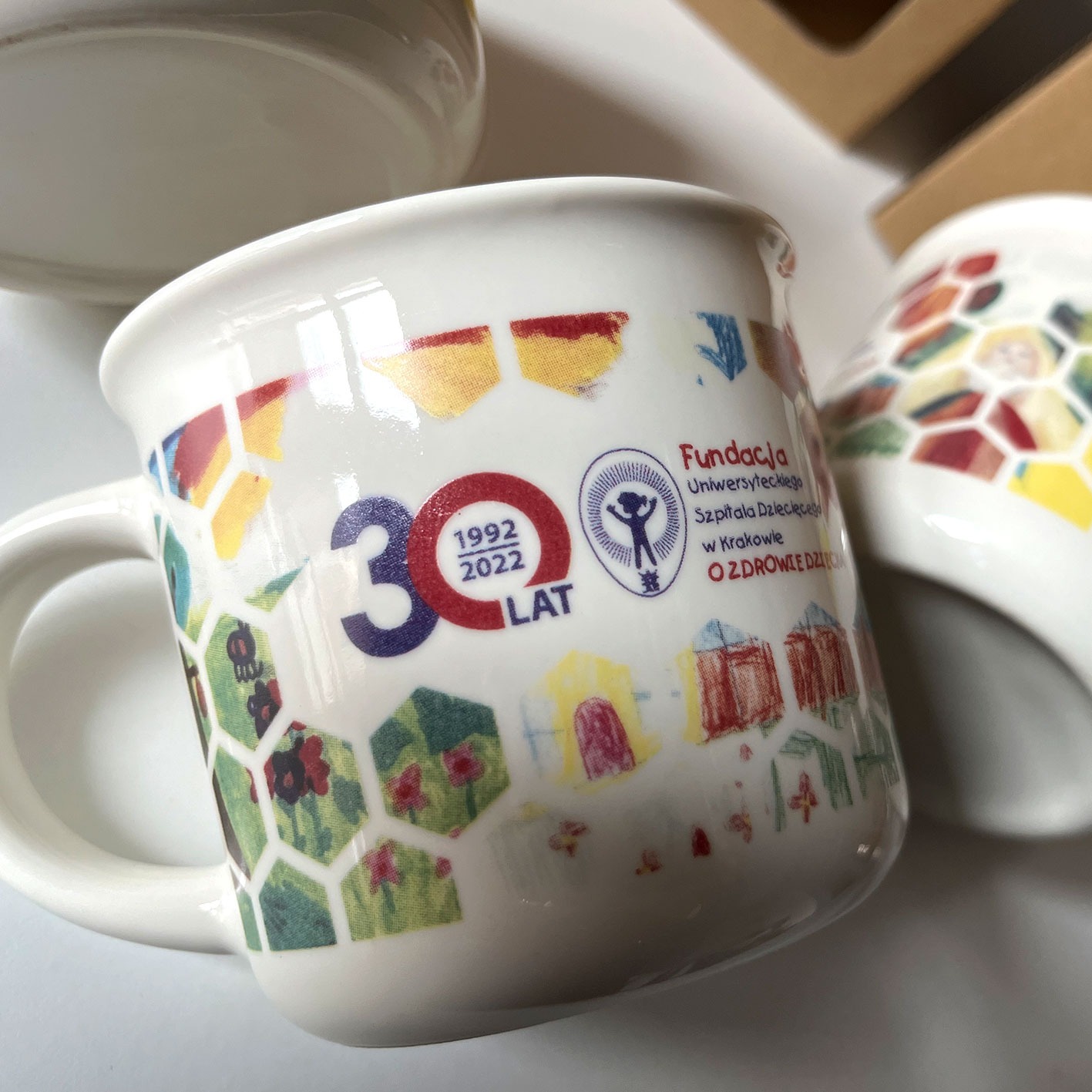 Dwa białe ceramiczne kubki z kolorowym nadrukiem motywów dziecięcych i logo fundacji, upamiętniające 30-lecie działalności szpitala dziecięcego w Krakowie, leżące na białym tle.