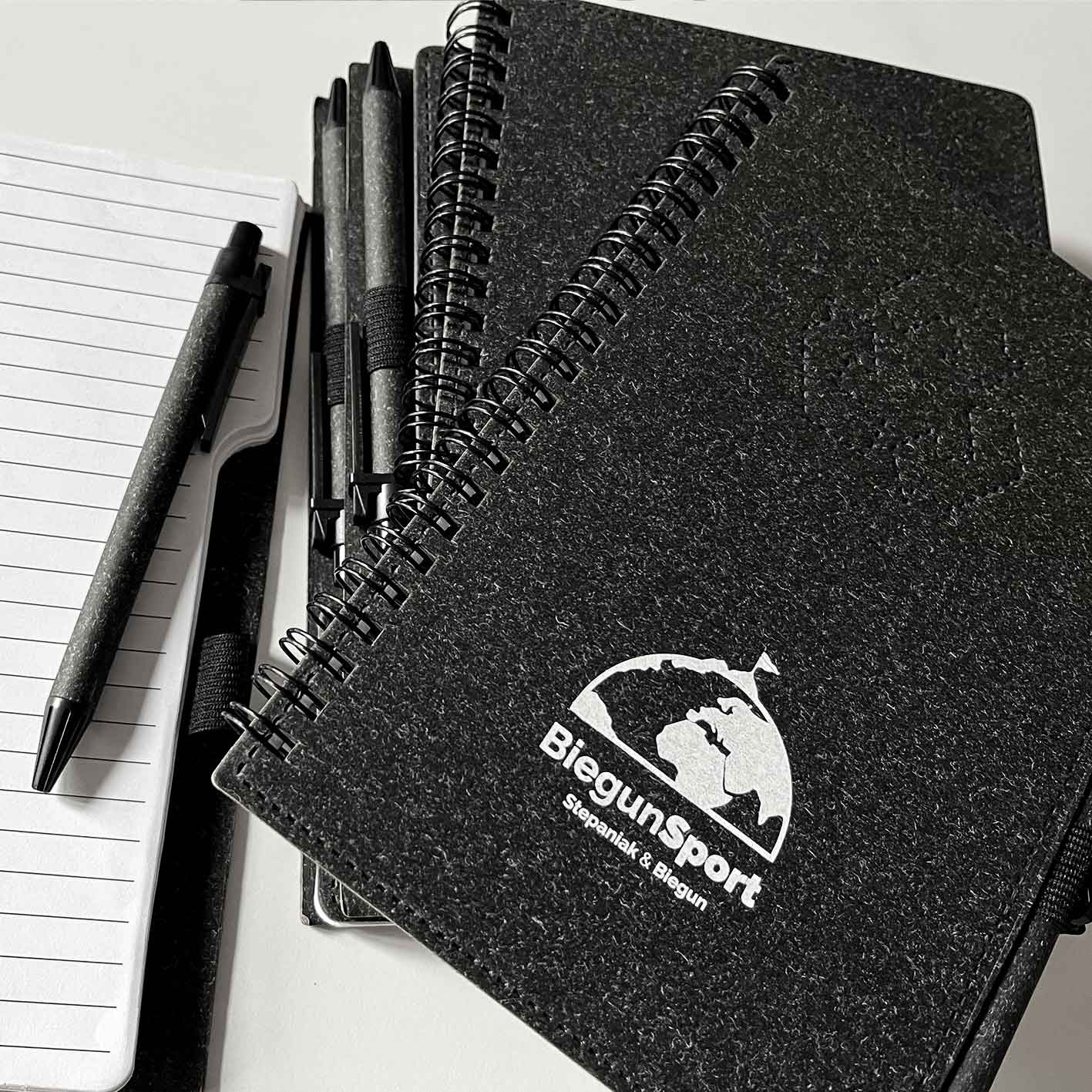 Stos czarnych notesów spiralnych z logo BiegunSport, obok otwarty notatnik w linie i czarny długopis z teksturą, ułożone na jasnym tle.