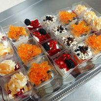 Taca z małymi, kwadratowymi deserami w przezroczystych pojemnikach: mini serniczki z owocami (jeżyny, porzeczki) i posypkami, niektóre z pomarańczową galaretką, inne z płatkami migdałów.