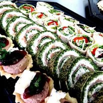 Aranżacja przekąsek na czarnej tacy: roladki szpinakowe z nadzieniem, jasne roladki z warzywami i mini tartaletki z pasztetem i żurawiną.