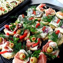 Różnorodne, ozdobne kanapeczki na czarnym, błyszczącym talerzu cateringowym, udekorowane świeżymi ziołami, oliwkami i papryką, widoczne także roladki z nadzieniem w tle.