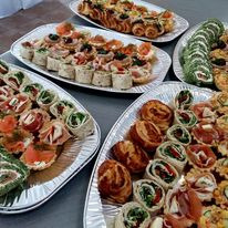 Różnorodne przekąski na srebrnych tacach, w tym roladki z zielonym szpinakiem, mini tartaletki z łososiem, tortille z warzywami i wędlinami oraz słone paluszki. Kompozycja na stole z szarego blatu.