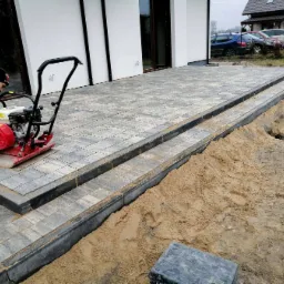 Pracownik zagęszcza wibratorem płytowym szarą kostkę brukową na tarasie przed białym domem z ciemnymi oknami, widoczny wykop z piaskiem.