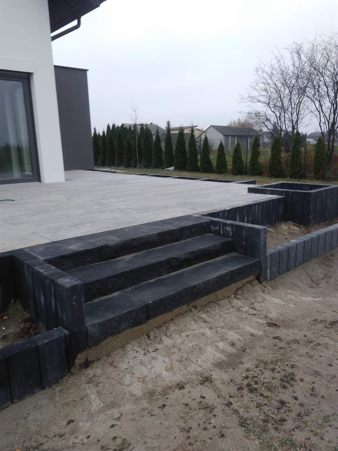 Schody z ciemnej kostki brukowej prowadzące z tarasu do ogrodu, widoczne obrzeża rabaty, tło stanowi nowoczesny dom i rząd tuj.