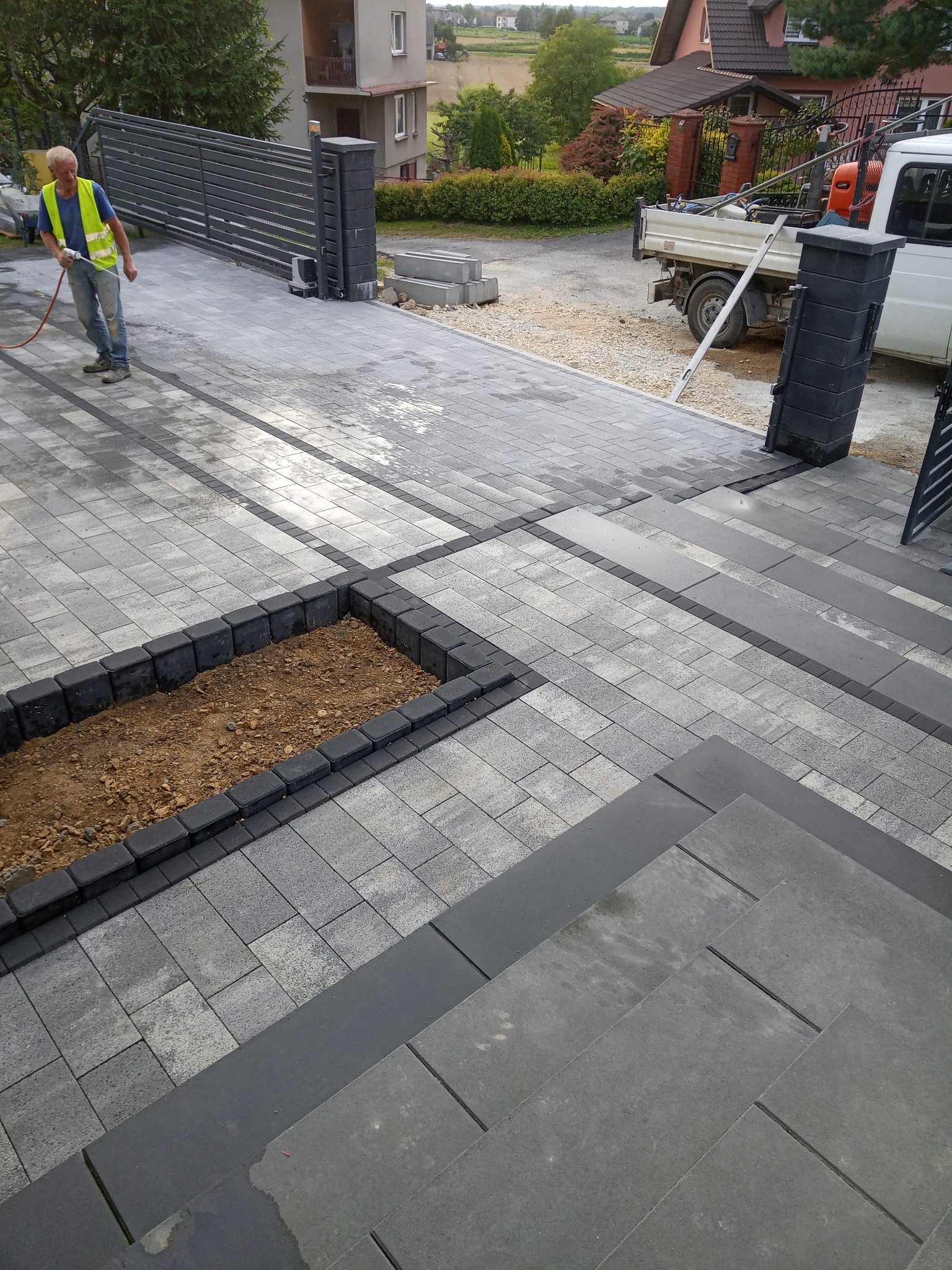 Ułożona szara kostka brukowa z czarnymi obrzeżami, klomb z ziemią, pracownik w odblaskowej kamizelce myjący kostkę, w tle brama wjazdowa i samochód dostawczy.