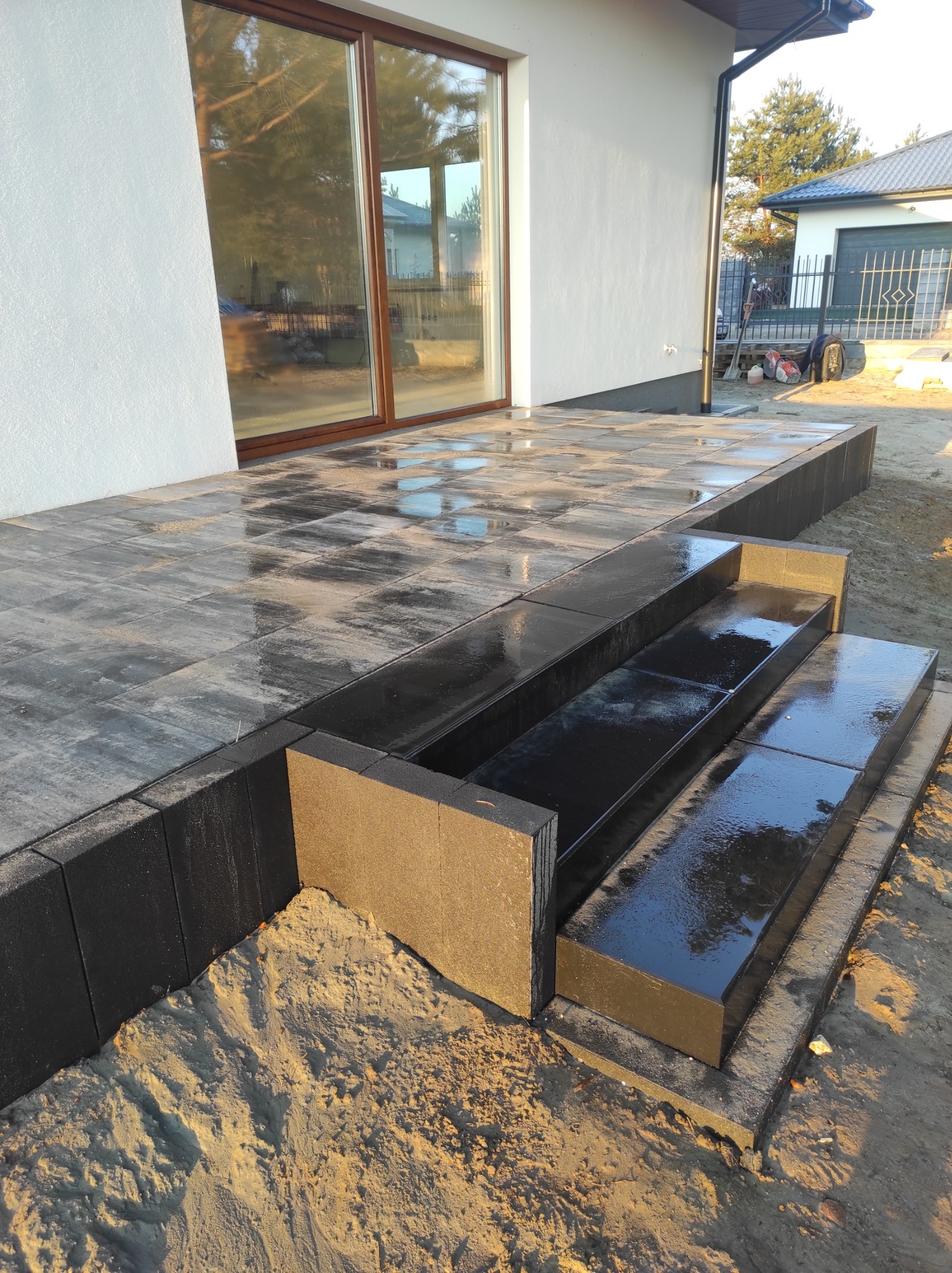 Nowoczesne schody z ciemnej kostki brukowej prowadzące do tarasu przy domu, świeżo po deszczu, z widocznym piaskiem i elementami ogrodu w tle.