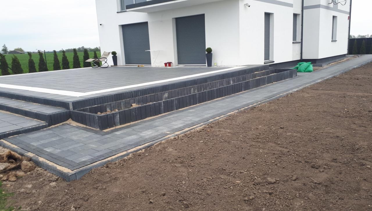 Nowoczesny taras z szarej kostki brukowej z białymi obrzeżami, schody z kostki prowadzące do domu z roletami zewnętrznymi, widoczne worki z zielonym wypełnieniem i trawnik w tle.