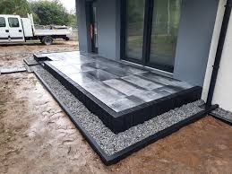 Nowoczesny taras z szarych płyt ułożonych na podwyższeniu z obramowaniem z czarnych elementów, wysypany drobnym szarym żwirem, z widocznym fragmentem elewacji budynku i samochodu dostawczego w tle.