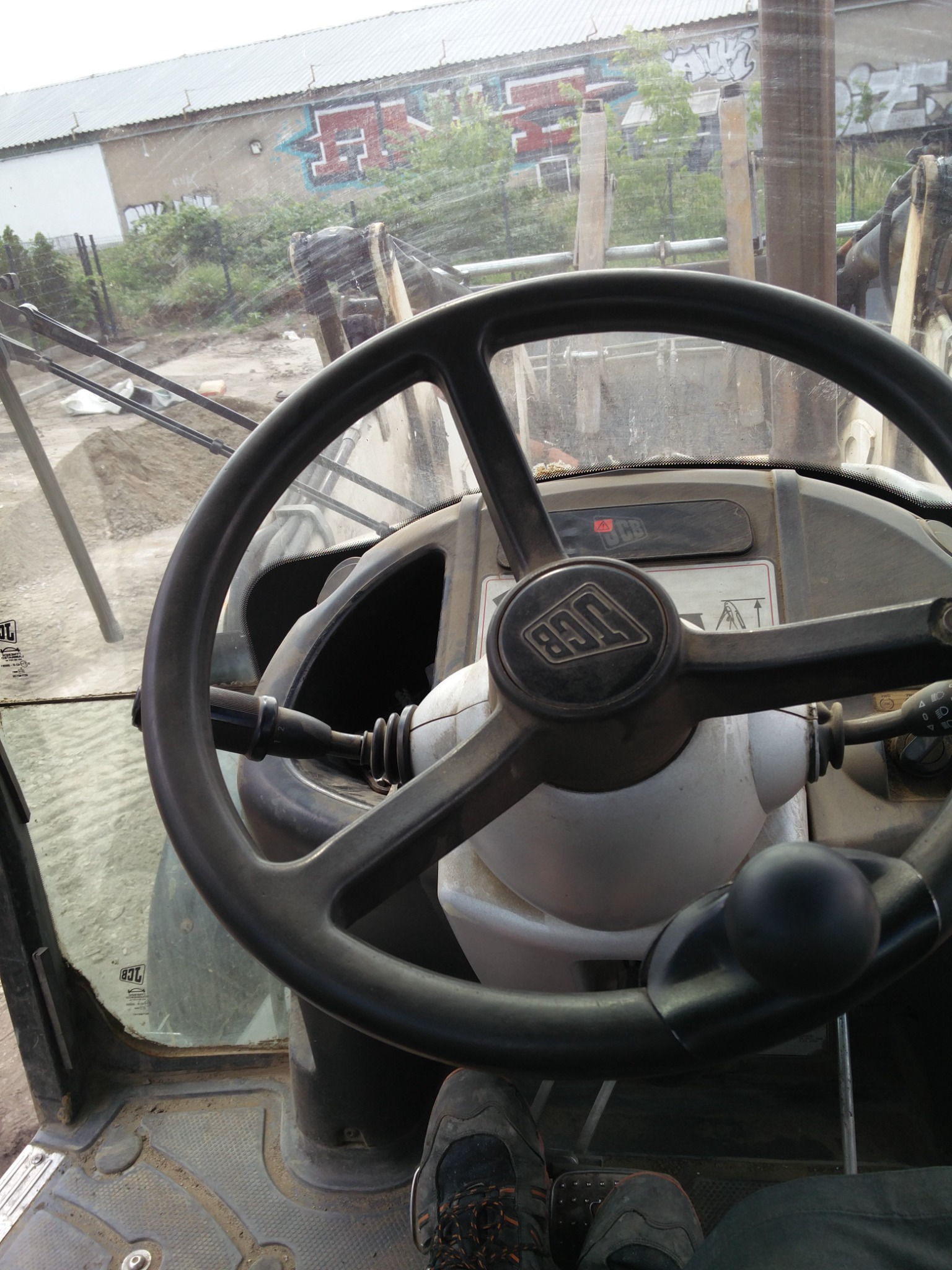 Wnętrze kabiny koparko-ładowarki JCB z widoczną kierownicą, deską rozdzielczą i fragmentem buta operatora, na zewnątrz plac budowy z hałdą ziemi i budynkiem z graffiti.