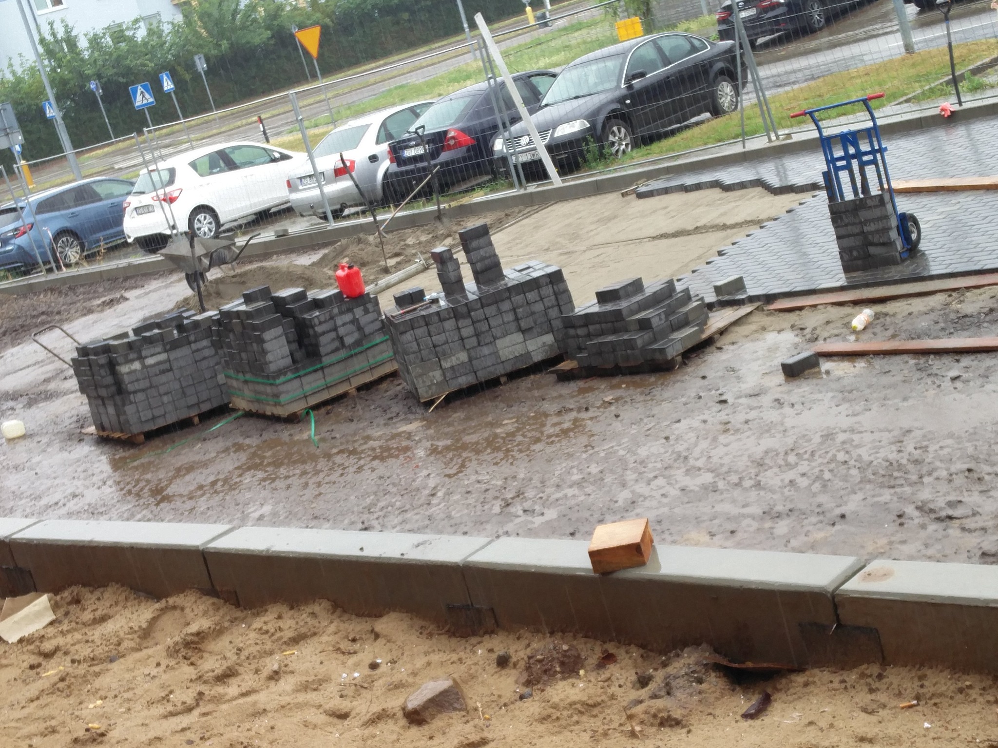 Stosy szarych kostek brukowych ułożone na paletach i w wózku transportowym na placu budowy, widoczne krawężniki i przygotowane podłoże, w tle zaparkowane samochody i znaki drogowe.