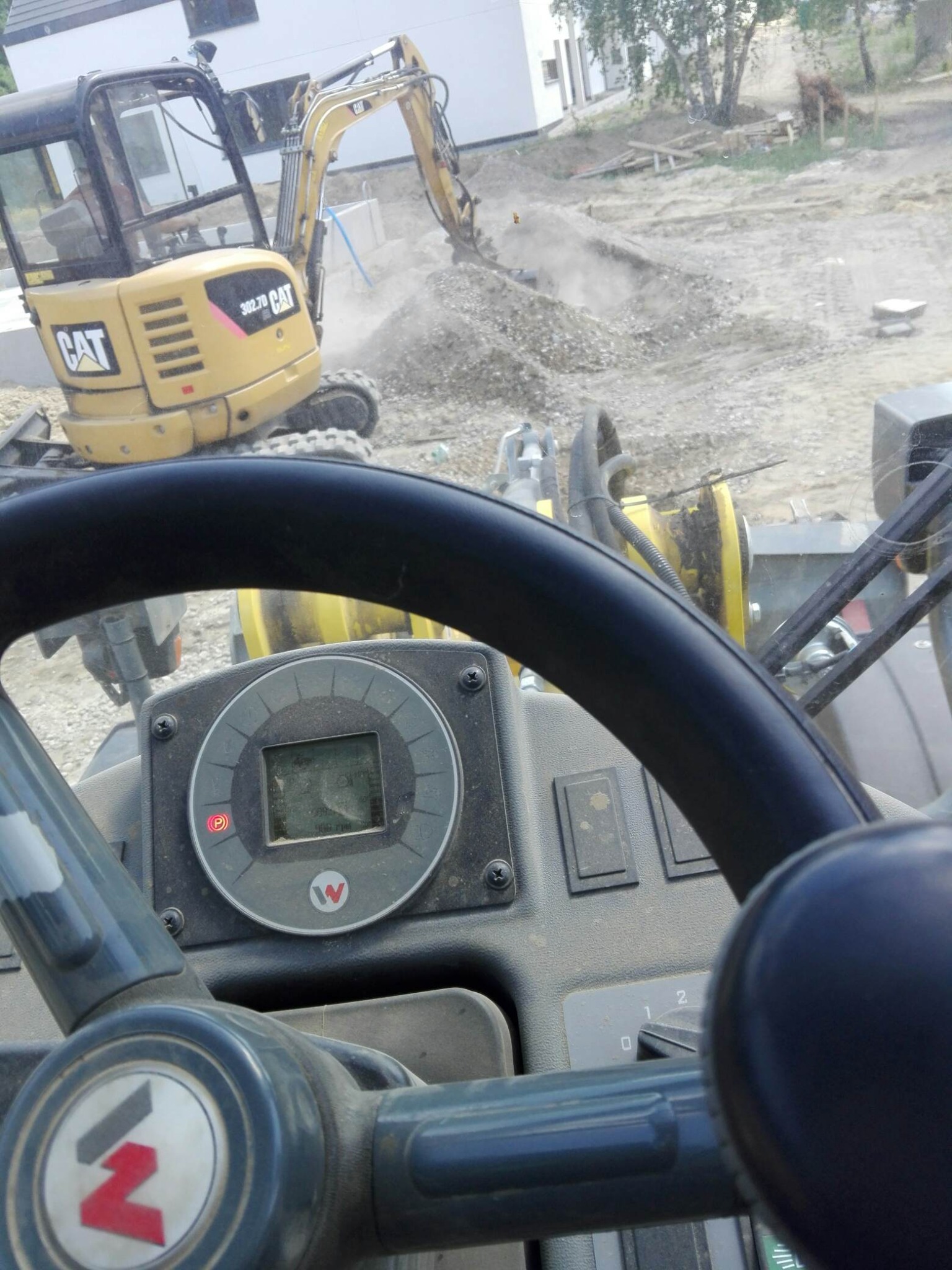 Widok z kabiny walca drogowego na koparkę CAT podczas prac ziemnych na placu budowy. W tle widoczny częściowo budynek w stanie surowym i hałda ziemi.