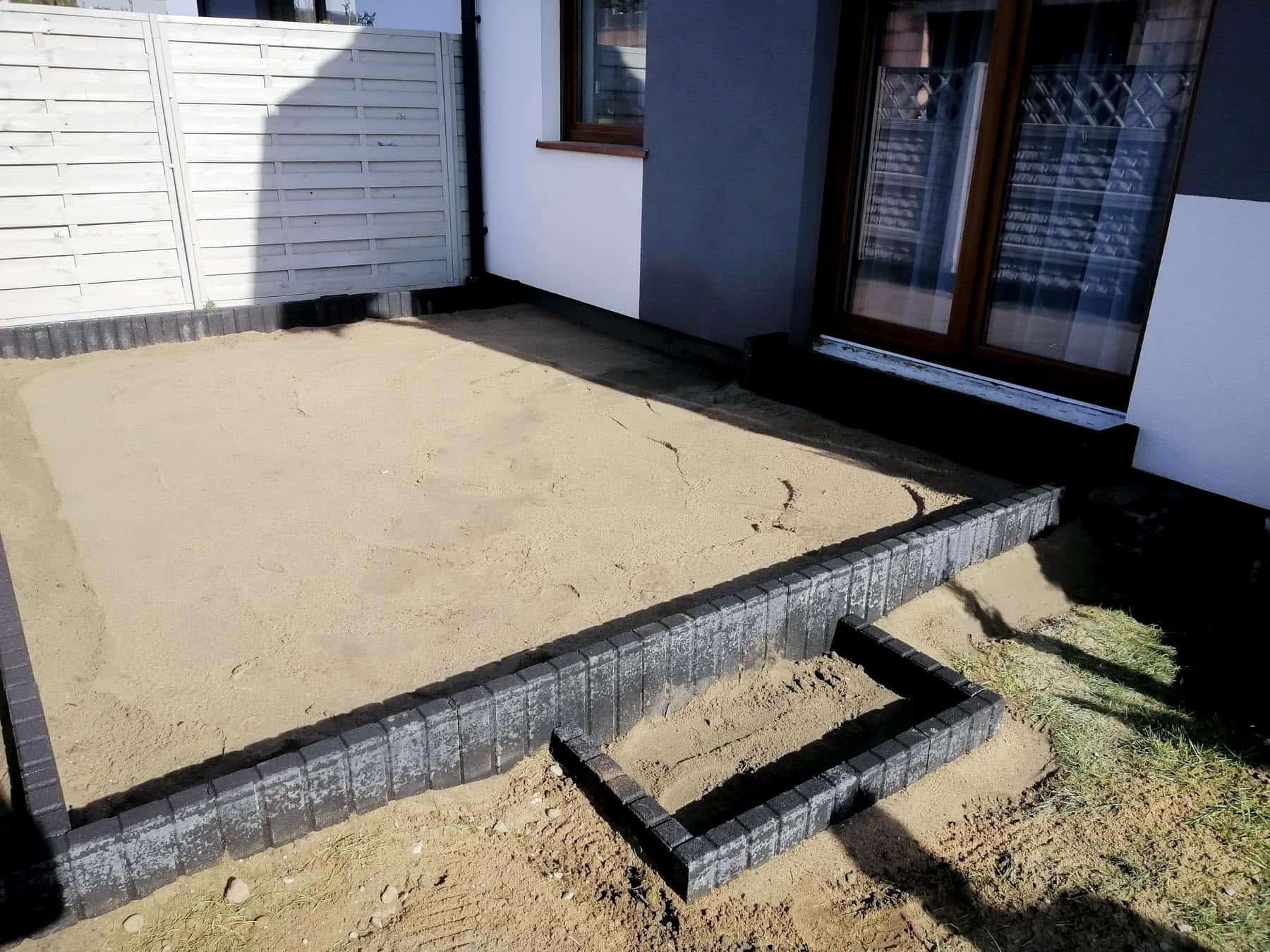 Przygotowanie podłoża pod taras: piasek wyrównany, obrzeża z betonowej kostki ułożone wokół przyszłego tarasu, fragment obrzeża ułożony w formie litery L, biały płot w tle.