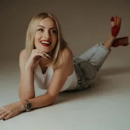 Portret uśmiechniętej blondynki leżącej na podłodze w dżinsach i białym topie, z czerwoną szminką i zegarkiem na ręce, z uniesionymi nogami w czerwonych butach.