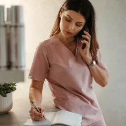 Kobieta w różowym uniformie lekarskim rozmawia przez telefon i jednocześnie pisze w otwartym kalendarzu na blacie, z widocznym zegarkiem na ręce.