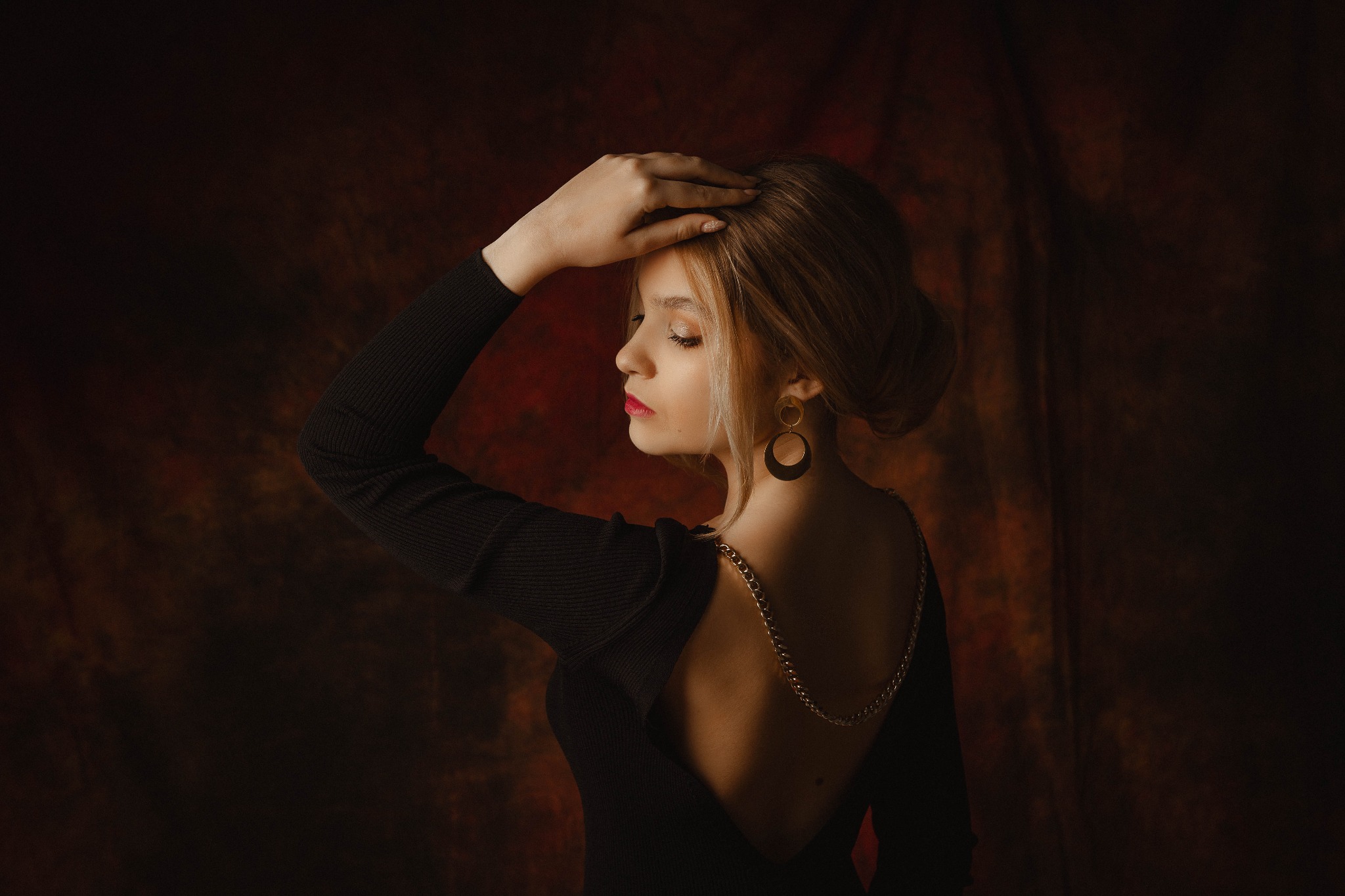 Portret kobiety z profilu, w czarnej sukience z odkrytymi plecami i złotym łańcuchem, pozującej na tle abstrakcyjnej, ciemnej tkaniny.