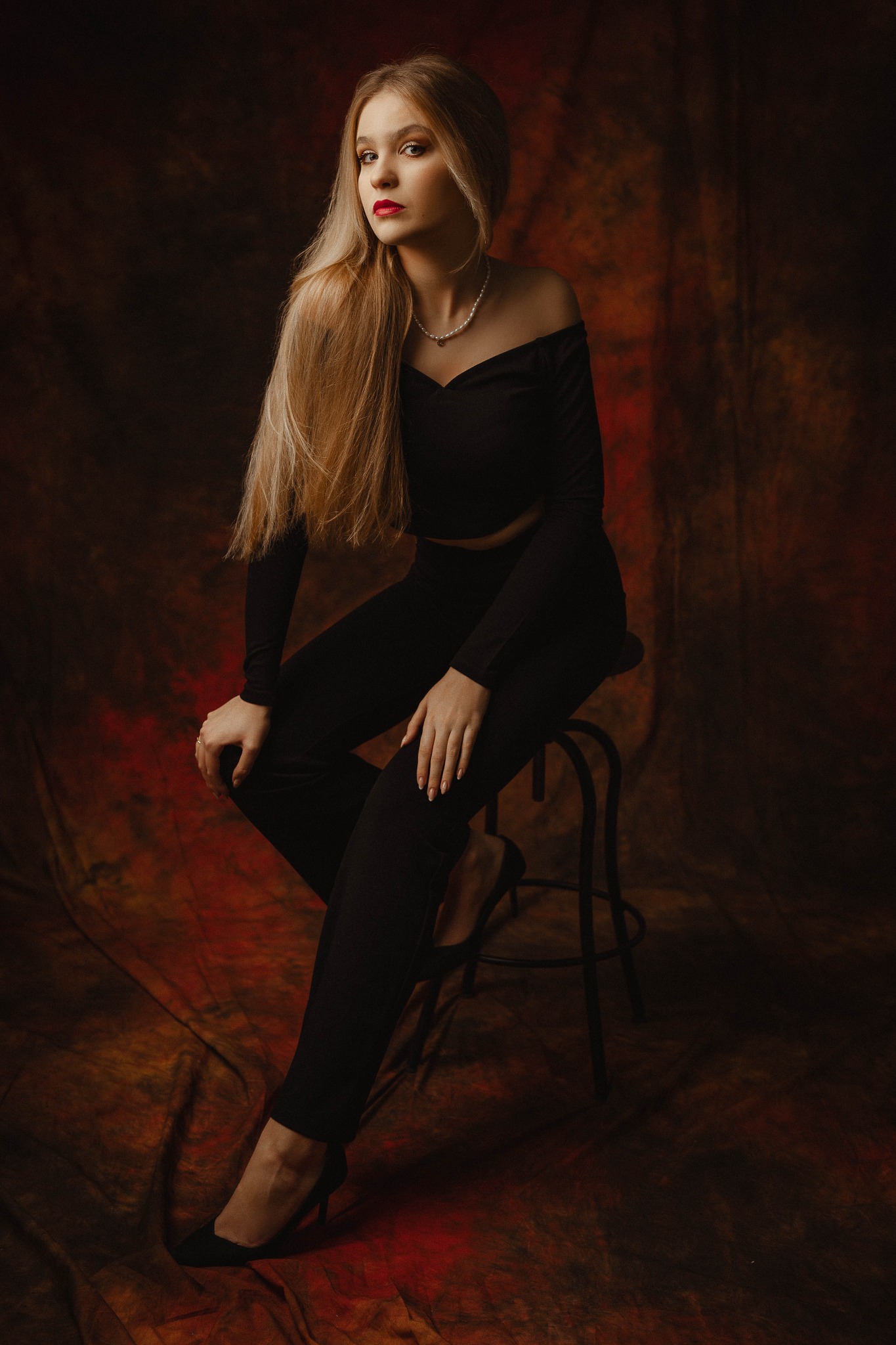 Portret kobiety w czarnym stroju siedzącej na stołku na tle abstrakcyjnego, ciemnego tła z czerwonymi tonami, z długimi blond włosami i perłowym naszyjnikiem.