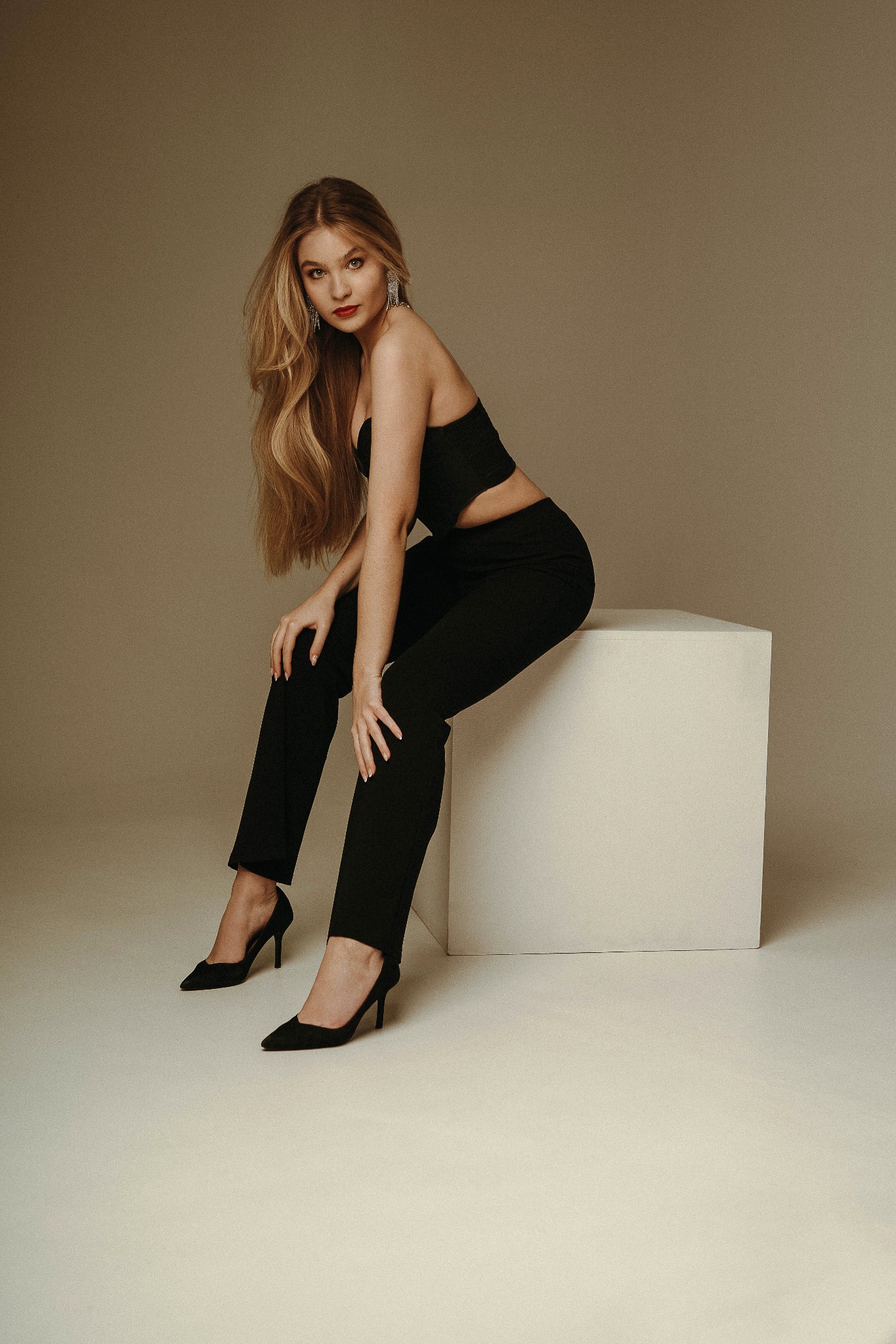 Portret kobiety w czarnym stroju siedzącej na białym sześcianie, z długimi blond włosami i kolczykami z frędzlami, na neutralnym tle.