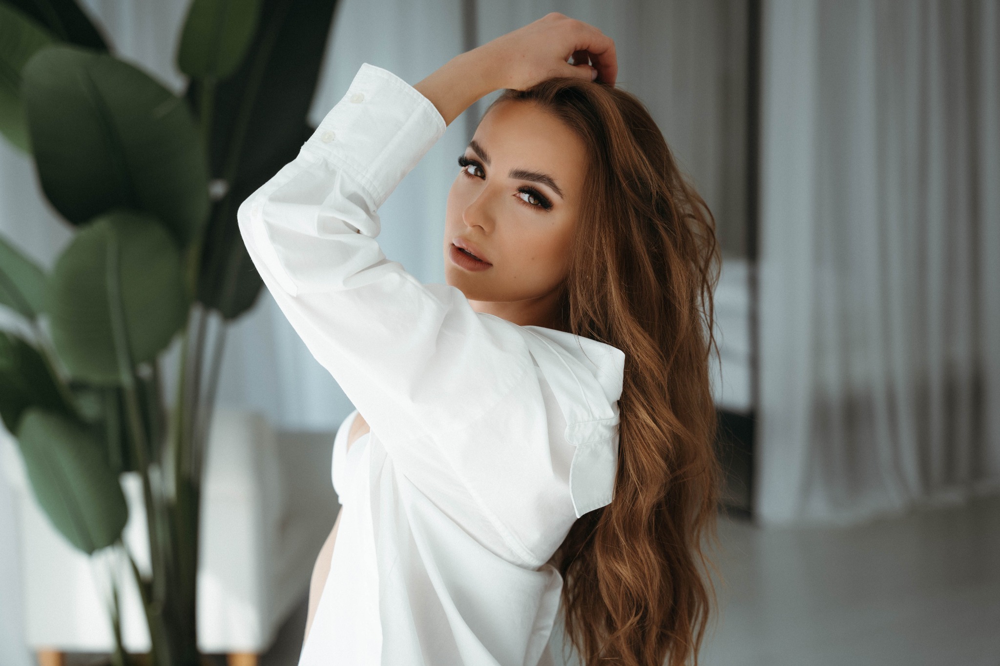 Portret kobiety w białej koszuli, z ręką we włosach, patrzącej przez ramię na tle zielonej rośliny doniczkowej i szarej zasłony.