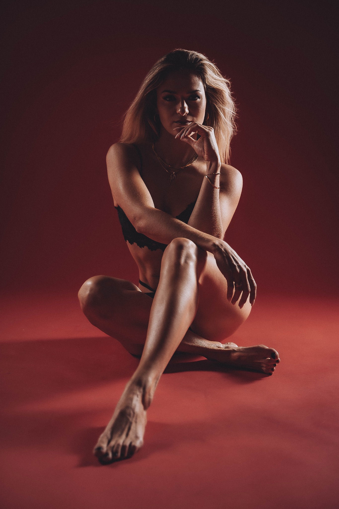 Portret kobiety siedzącej na czerwonym tle, w czarnej bieliźnie, z ręką podpartą pod brodą, spoglądającej w obiektyw z lekkim cieniem na twarzy.